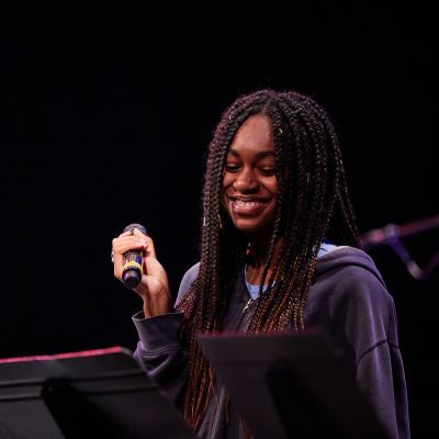 Woman on stage holding microphone smiling and looking down at music on stand